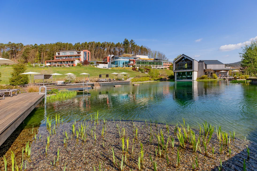 Hotelgebäude und Steg am natürlichen Schwimmteich mit grünen Pflanzen unter klarem Himmel