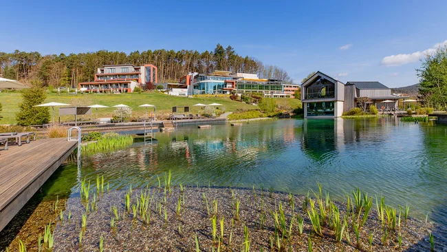 Hotelgebäude und Steg am natürlichen Schwimmteich mit grünen Pflanzen unter klarem Himmel