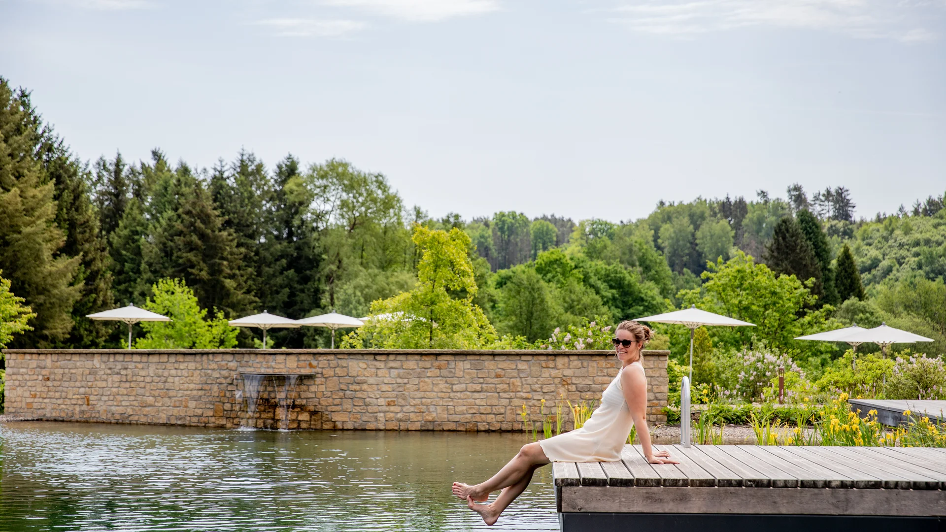 Pressebilder Frau sitzt entspannt am Steg über einem Teich mit Garten und Bäumen im Hintergrund