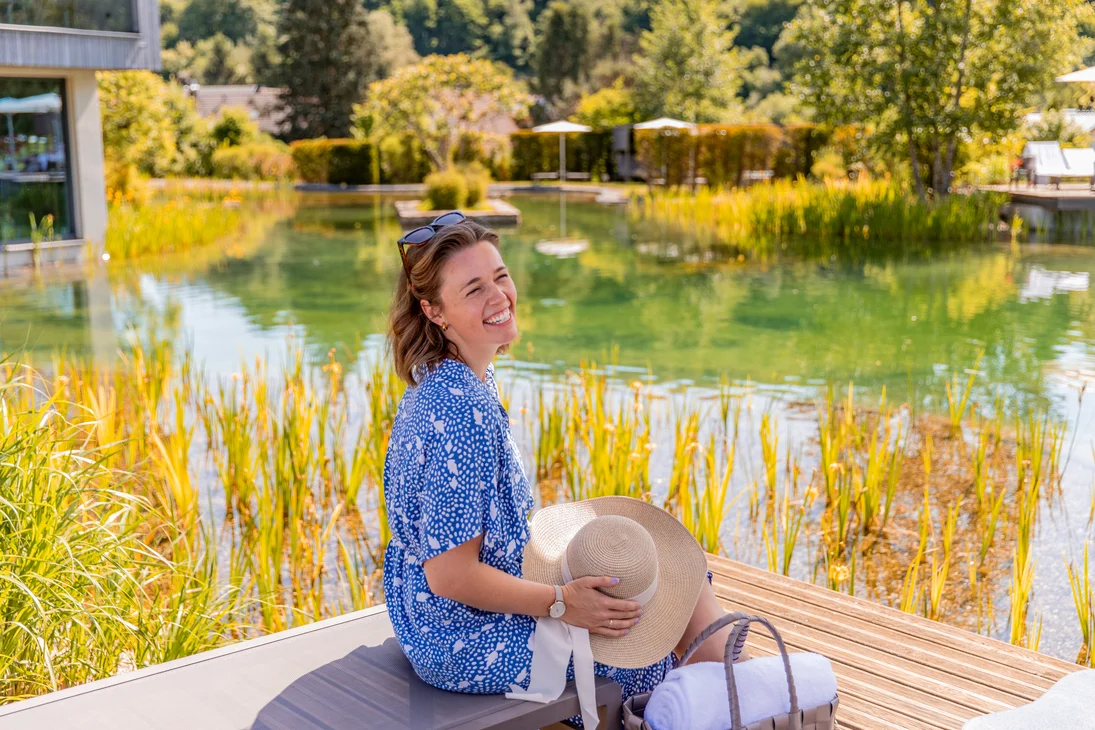 Ihr Hotel im Dahner Felsenland: Pfalzblick Wald Spa Resort Frau in blauem Kleid sitzt lächelnd am Teich mit Sonnenhut und Tasche