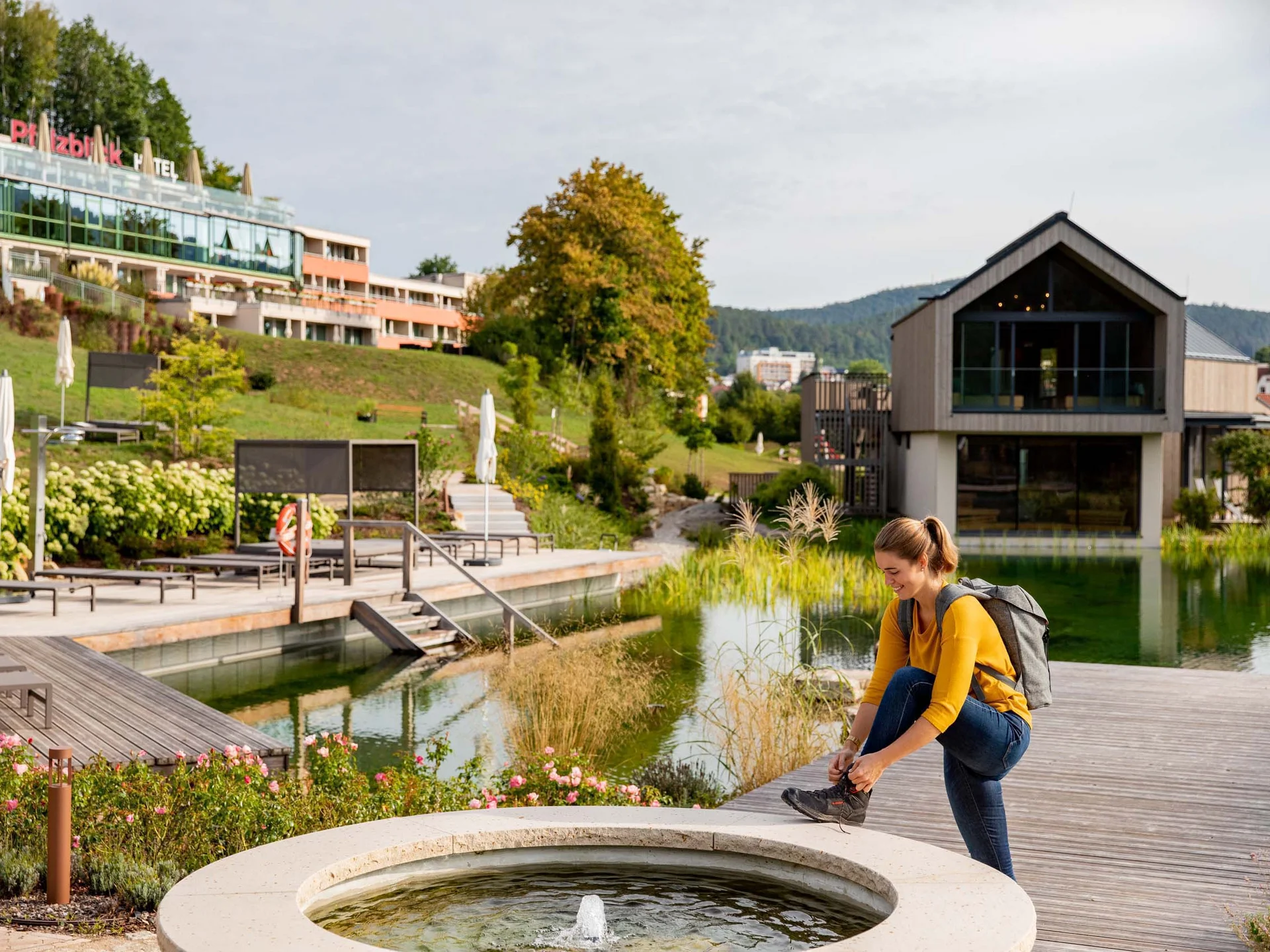 Frau bindet Wanderschuh am Teich mit Hotel und Natur im Hintergrund