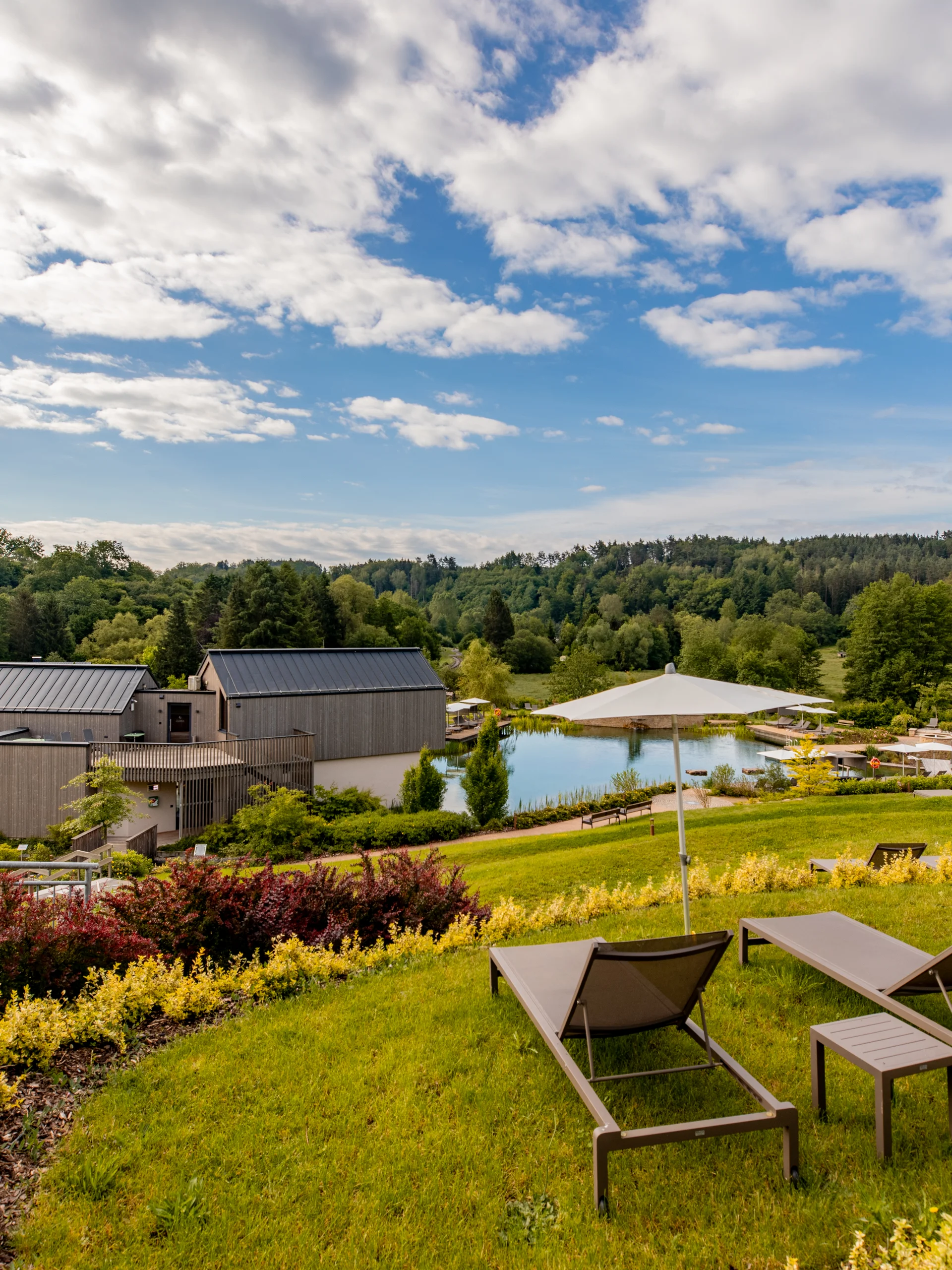 Liegestühle mit Sonnenschirmen vor Teich und Häusern in grüner Landschaft