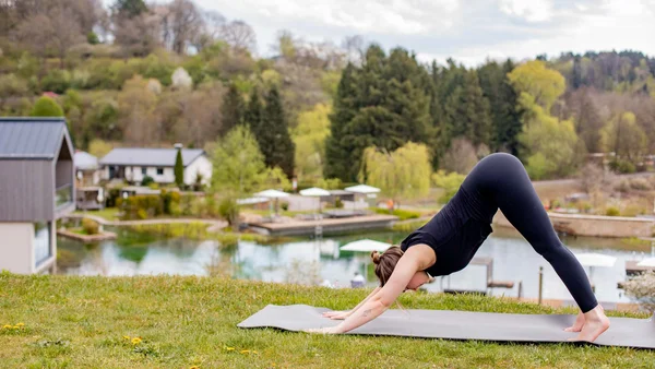 Frau macht Yogaübung auf Matte im Freien mit Teich und Häusern im Hintergrund