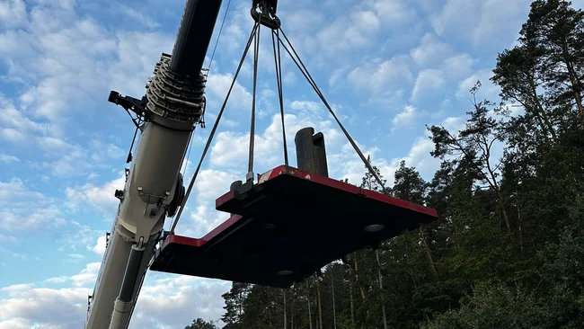 Une grue mobile soulève une grande plateforme noire près d’une forêt