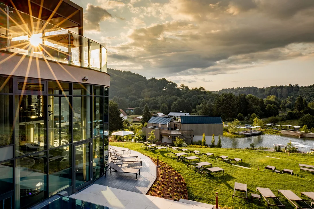 Sonnenuntergang über modernem Gebäude und Liegestühlen am See in grüner Landschaft