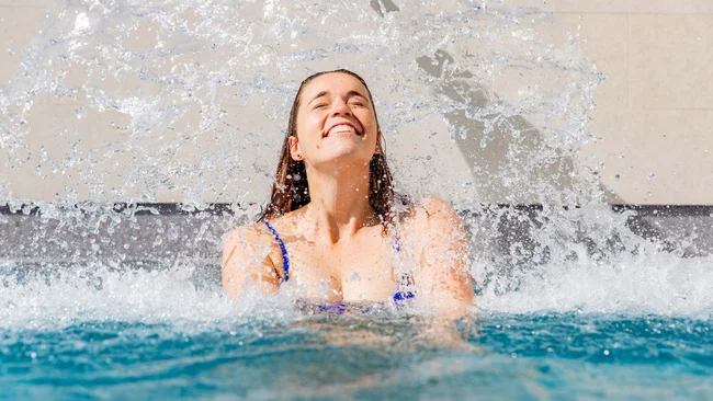 Frau in blauem Badeanzug springt aus Wasser mit Spritzwasser um sie herum