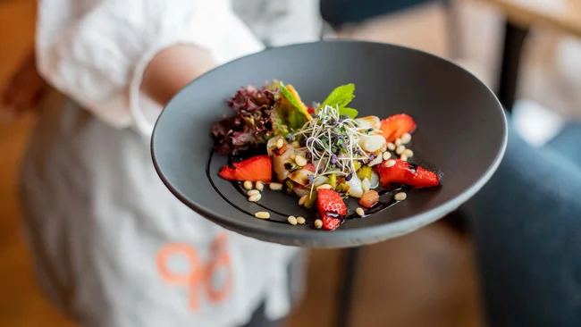 Person hält schwarzen Teller mit Salat, Erdbeeren, Samen und grünen Kräutern