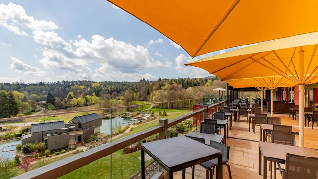 Sonnige Terrasse mit Tischen und Stühlen mit Ausblick auf See und bewaldete Hügel